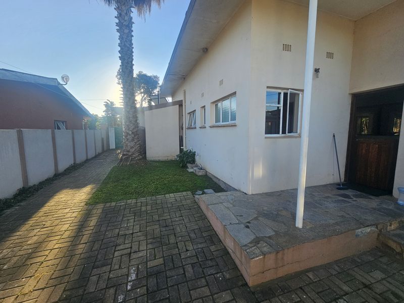 Rear exterior view showing a paved driveway, stone patio area, and boundary wall, with a side entrance to the house.
