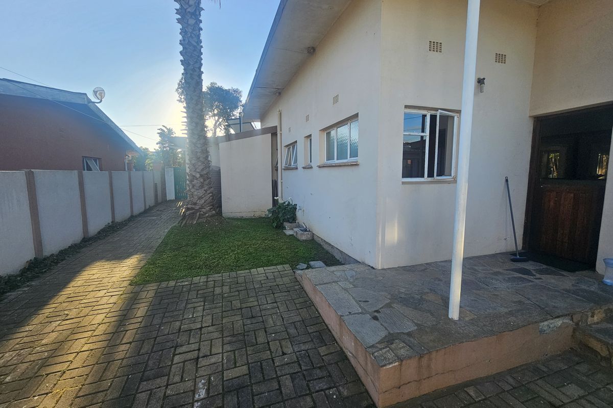 Rear exterior view showing a paved driveway, stone patio area, and boundary wall, with a side entrance to the house.