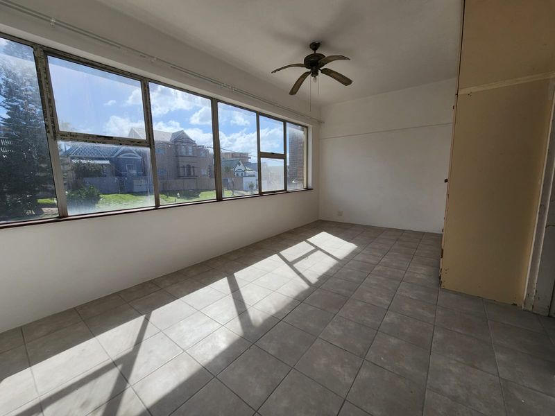 A sunlit, empty living area featuring large windows, tiled flooring, and a ceiling fan, offering a blank canvas for renovation.