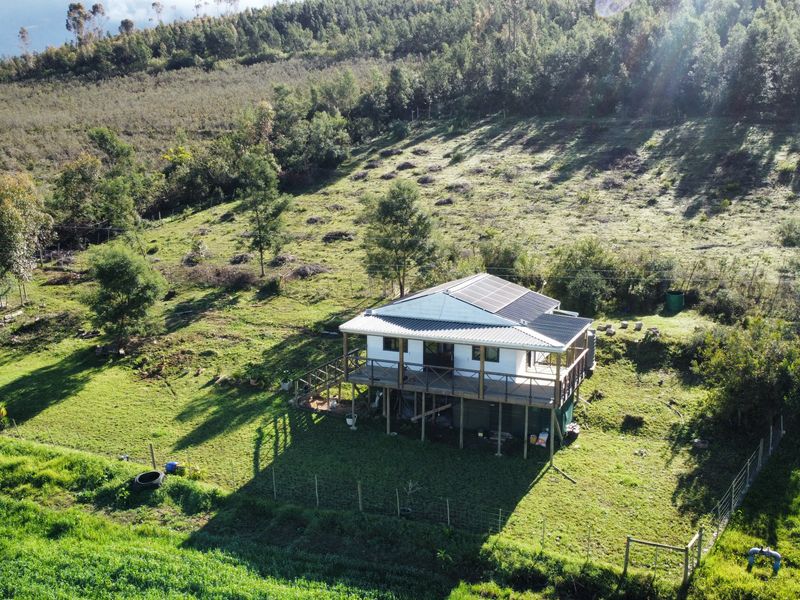 Aerial view of a rural property featuring a raised dwelling with a metal roof, solar panels, and an expansive timber deck set in a landscape.