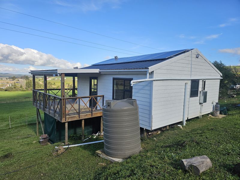 Rear view of a farm cottage featuring a raised wooden deck, solar panel installation, and a large water storage tank set in a rural landscape.