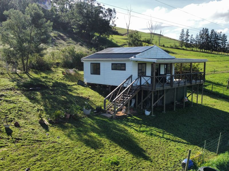 Elevated view of a rural farm cottage featuring a raised timber deck, metal roofing, and installed solar panels set within a grassy landscape.