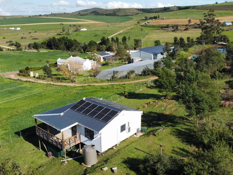Aerial view of a farm property featuring a main dwelling with solar panels, a timber deck, water tank, and surrounding agricultural land.