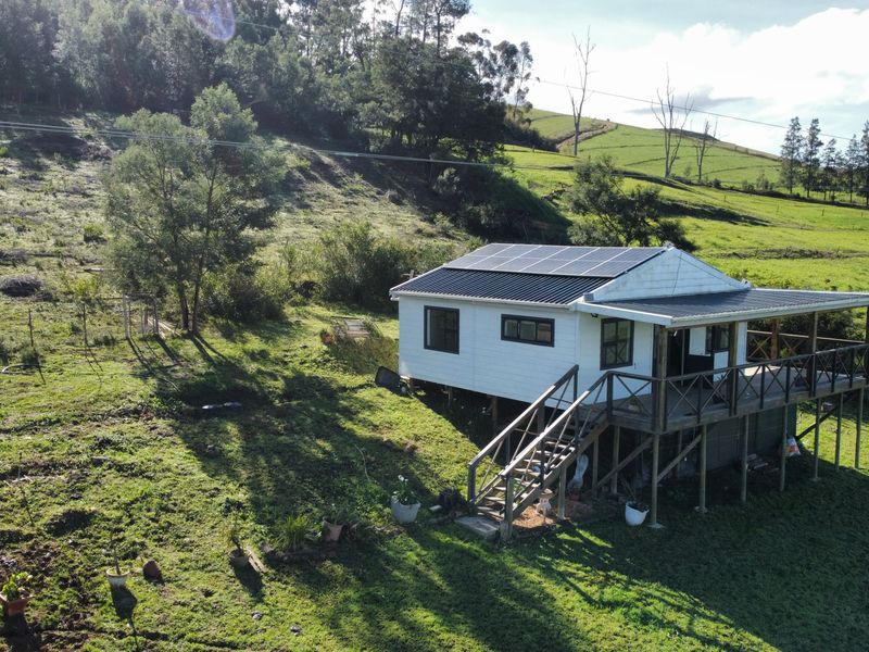 Aerial view of a rural farm cottage featuring a timber deck, staircase, and roof-mounted solar panels set within a rolling green landscape.