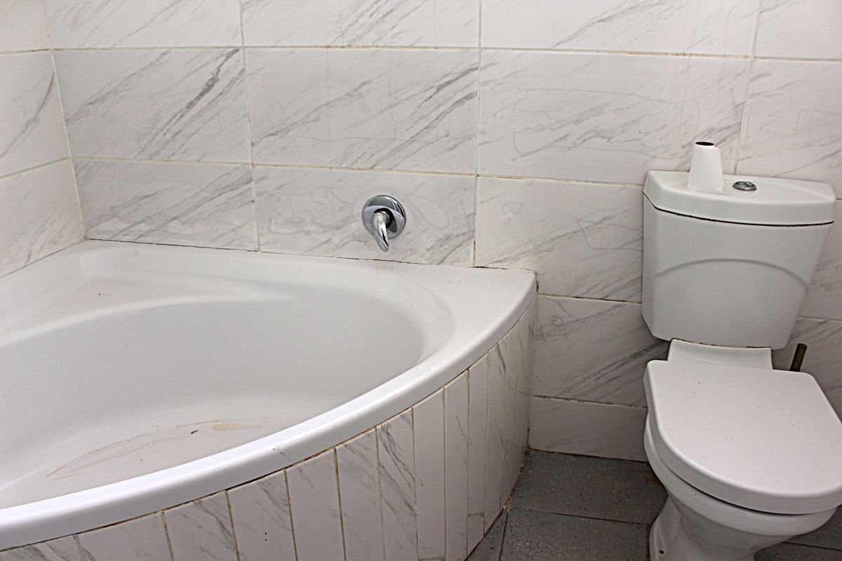 A compact bathroom featuring a corner bathtub and toilet with marble-look wall tiling, requiring some maintenance and cosmetic updates.