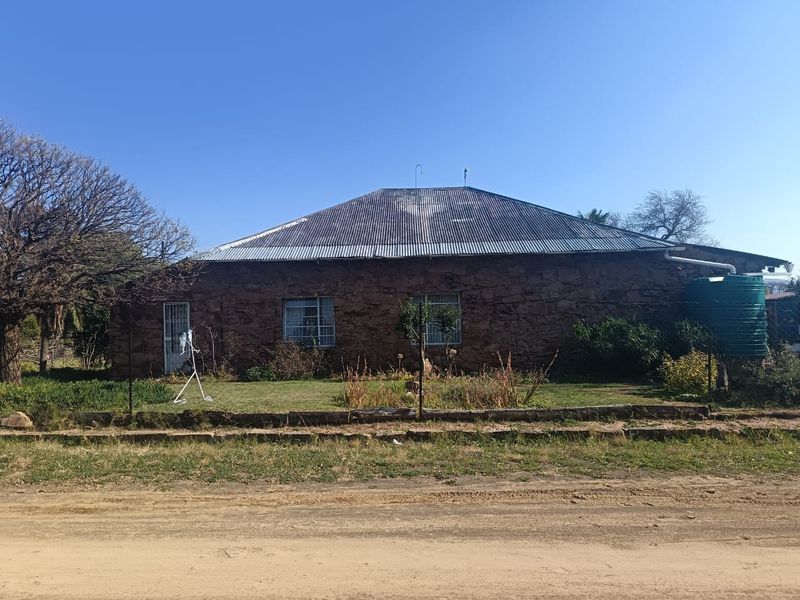 A rustic stone-walled house featuring a corrugated metal roof and an attached rainwater tank, situated on a dirt road.
