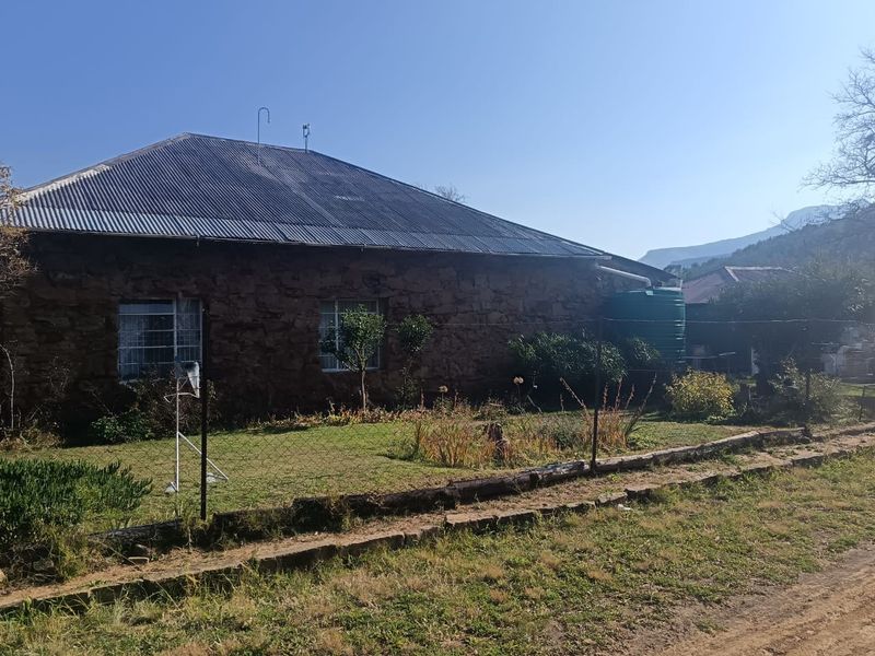 Rear view of a stone-walled house featuring a corrugated metal roof, guttering system, and an attached water storage tank set within a garden area.