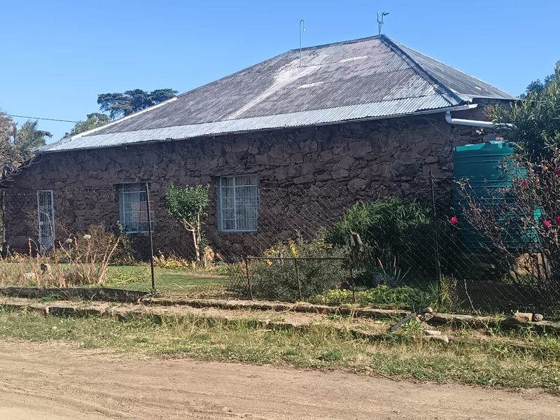 A rustic stone-walled house featuring a corrugated metal roof, security fencing, and an integrated water storage tank, situated along a dirt road.
