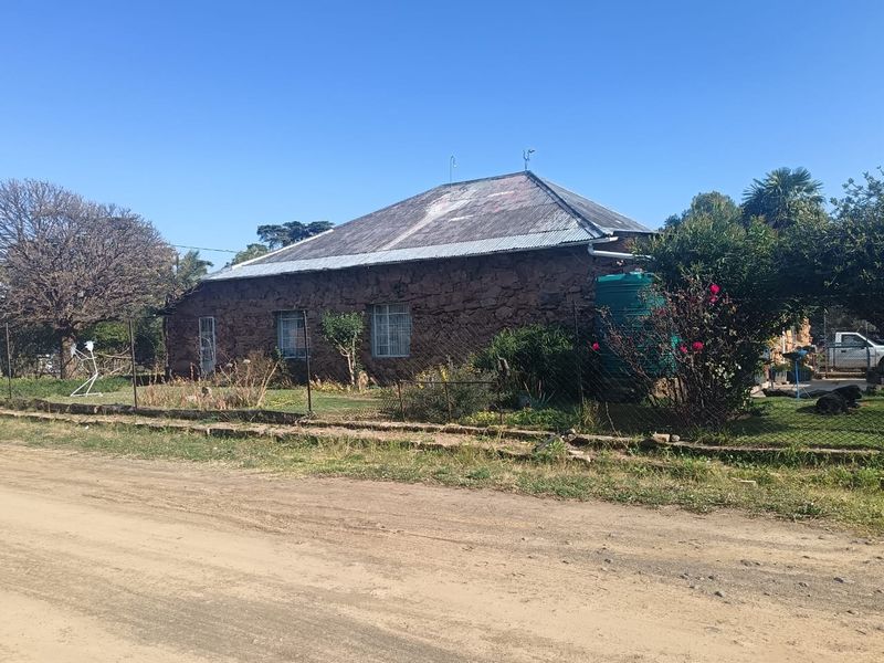 A rustic stone-walled house featuring a corrugated metal roof, security bars on windows, and an external water storage tank, situated along a dirt road.