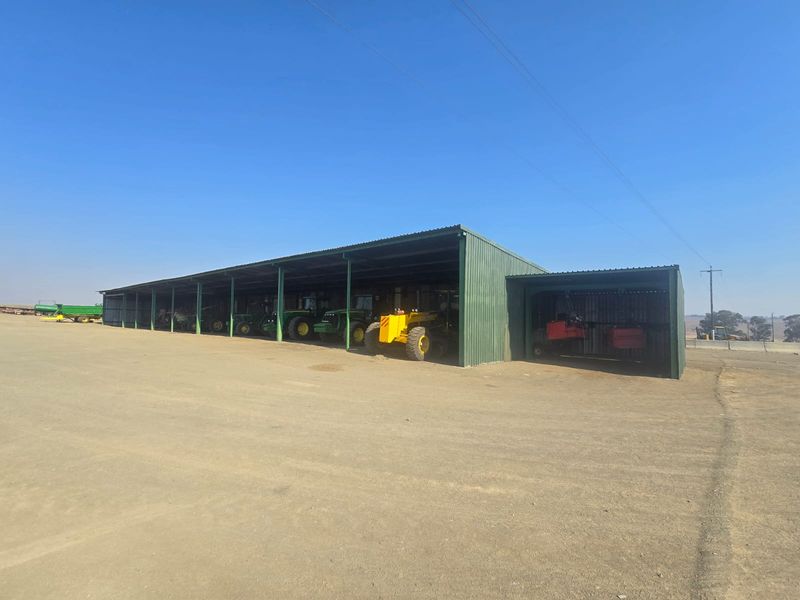 Large open-sided agricultural equipment shed with corrugated metal roofing and steel structural supports, suitable for farm machinery storage.