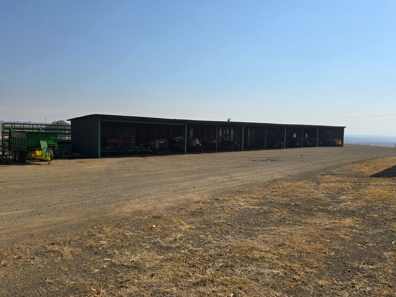 A large, open-sided agricultural machinery shed with a metal roof, providing covered storage for farm equipment on a gravel yard.