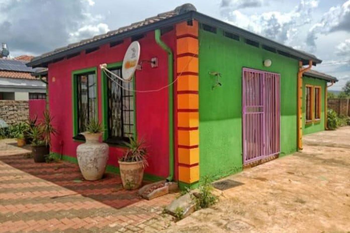 A detached house exterior featuring a vibrant, multi-colored facade with security gates and tiled roof, set on a paved and partially unpaved plot.