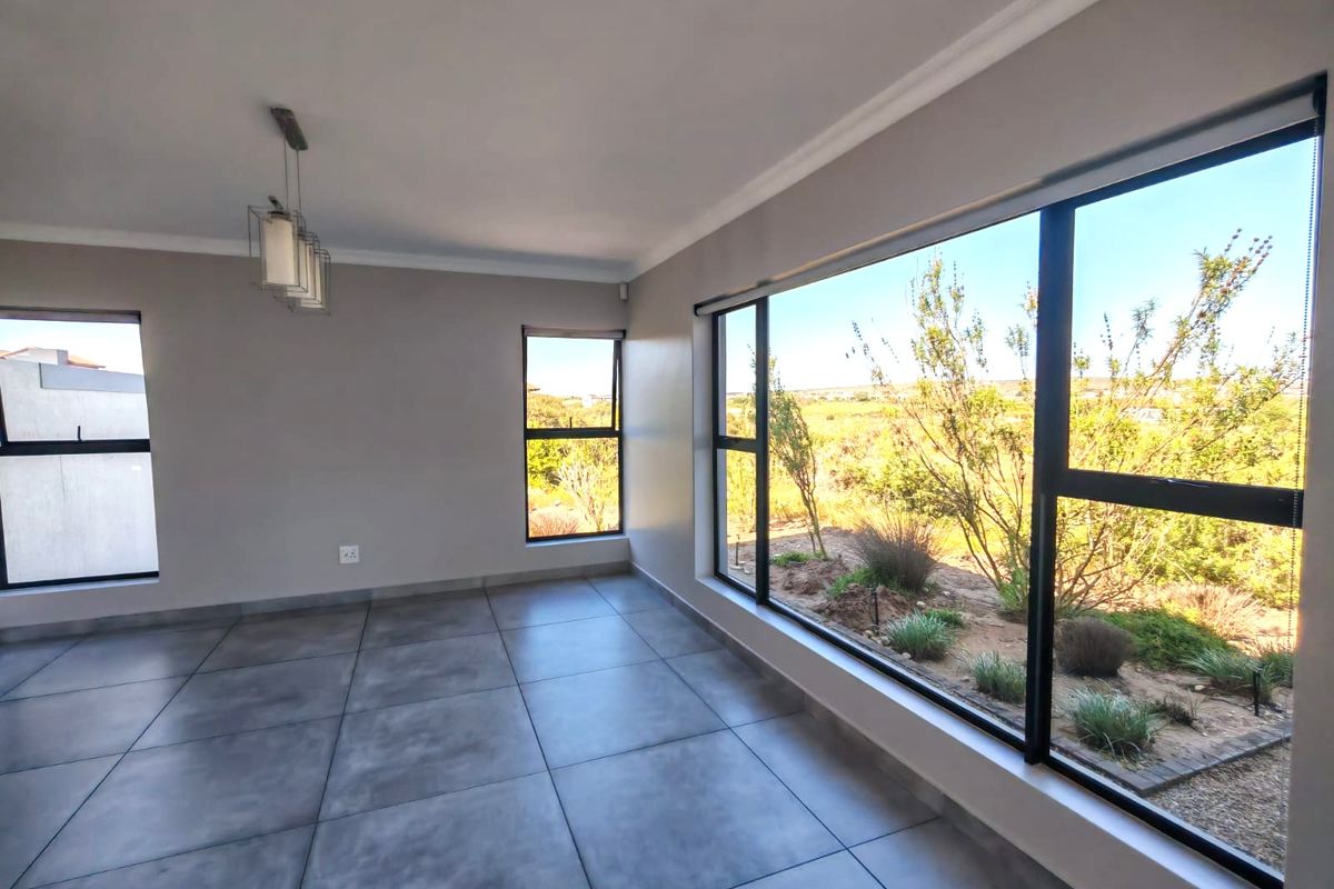 A bright, empty living area featuring large aluminium-framed windows with garden views, modern grey floor tiles, and a contemporary pendant light fixture.