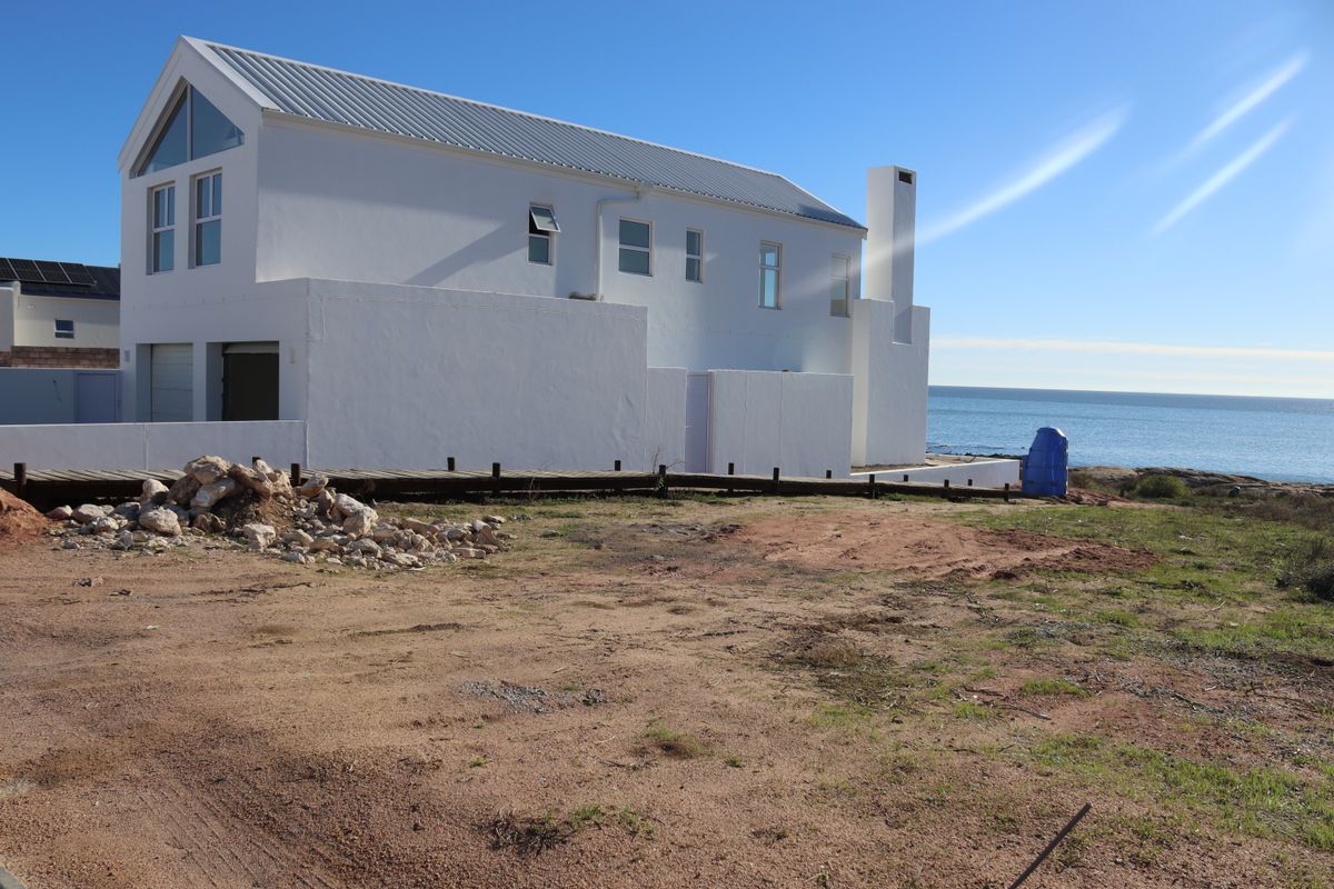 Rear view of a modern white coastal home featuring a metal roof, chimney, and timber decking, situated on an unlandscaped plot overlooking the ocean.