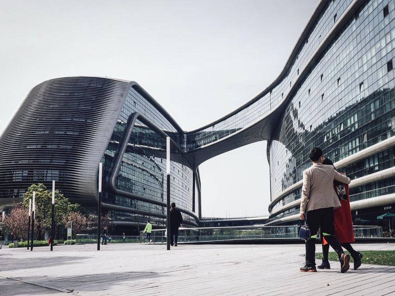Modern architectural commercial complex featuring curved glass facades and a connecting skybridge, set within a paved urban plaza.
