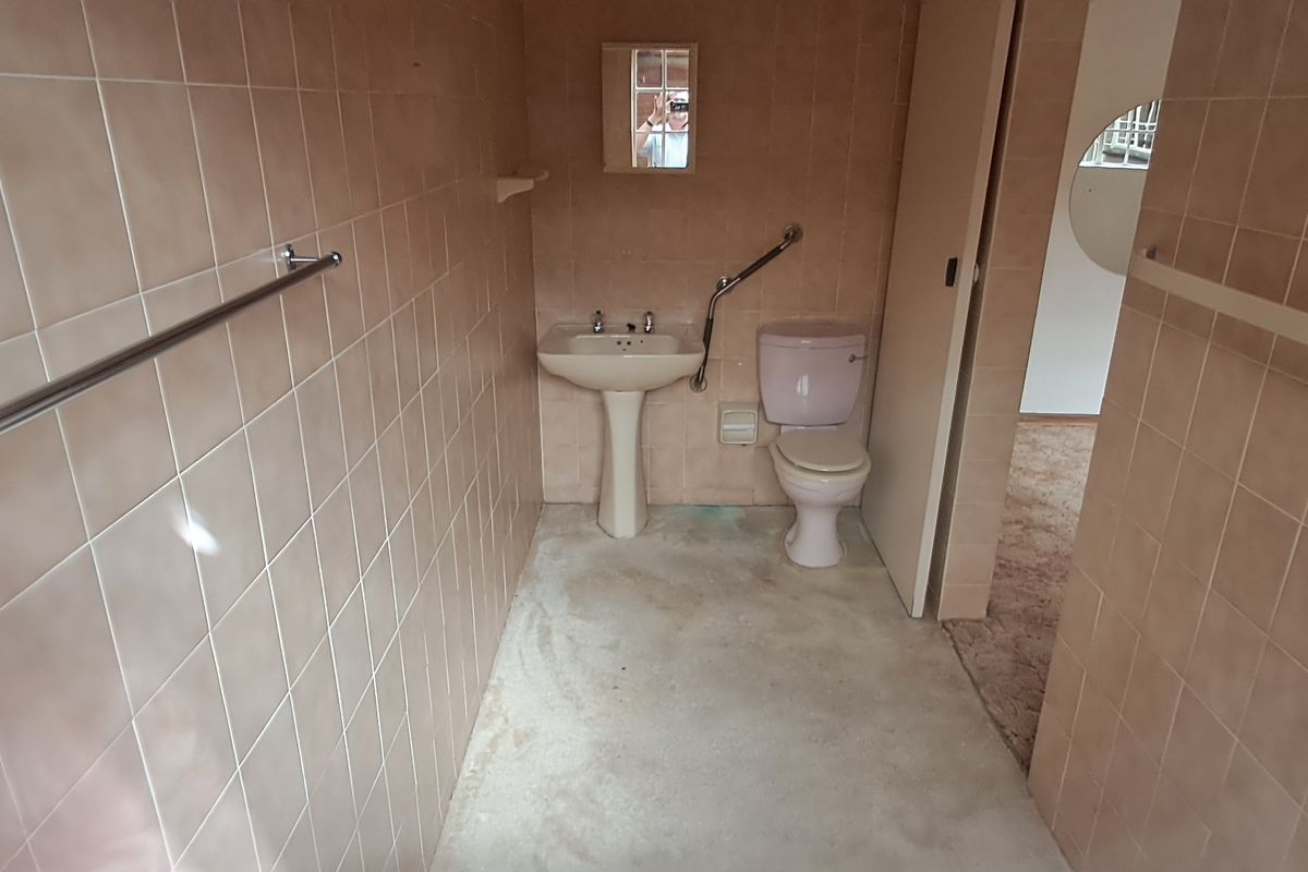 A dated bathroom featuring a pedestal basin, toilet, and grab rail, with full-height wall tiling and a worn floor surface.