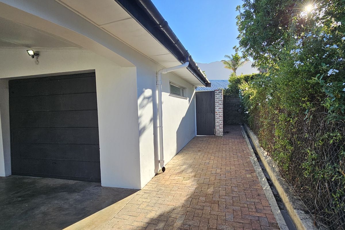 A paved side access path leading to a garage, featuring clean white exterior walls, guttering, and established boundary hedging.
