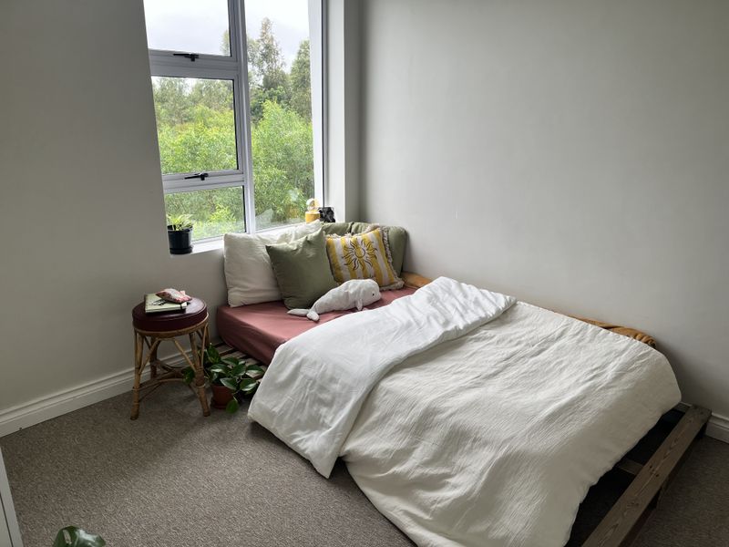 A bright bedroom featuring neutral walls, carpeted flooring, and a large window providing natural light and a pleasant green outlook.