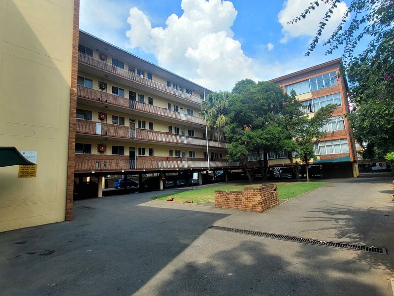 Exterior view of a multi-story apartment complex featuring brick facade, open-air balconies, and covered ground-level parking areas with a central paved courtyard.