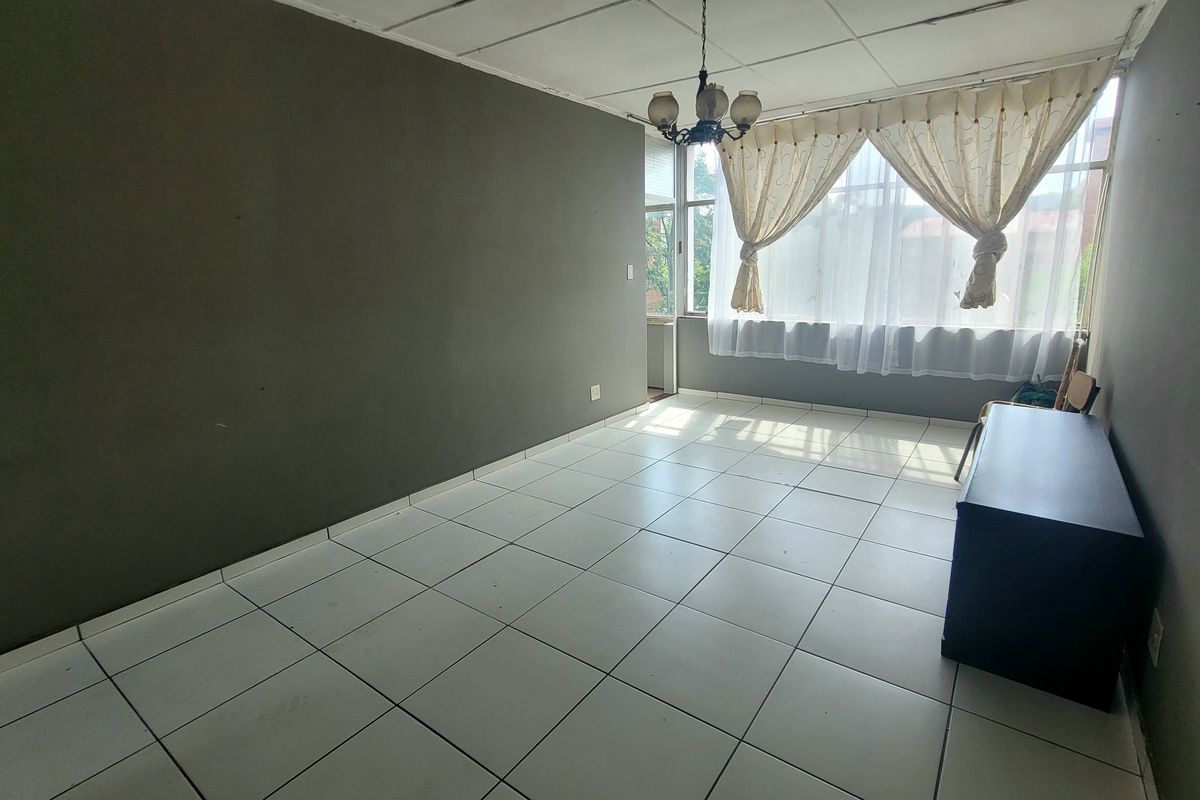 A spacious, empty living room featuring white tiled flooring, dark grey walls, and a large window with natural light.
