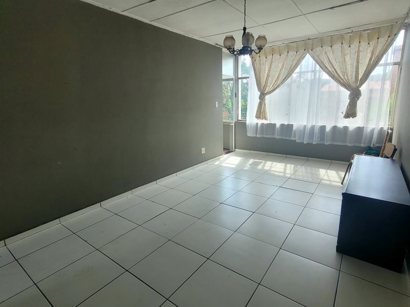 A spacious, empty living room featuring white tiled flooring, dark grey walls, and a large window with natural light.