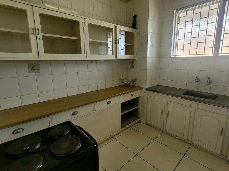 A dated kitchen featuring white cabinetry, tiled splashbacks, tiled flooring, and a stainless steel sink with security bars on the window.
