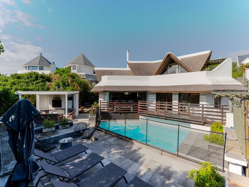 Outdoor entertainment area featuring a swimming pool with glass fencing, a covered patio with built-in braai, and a distinctive thatched roof design.
