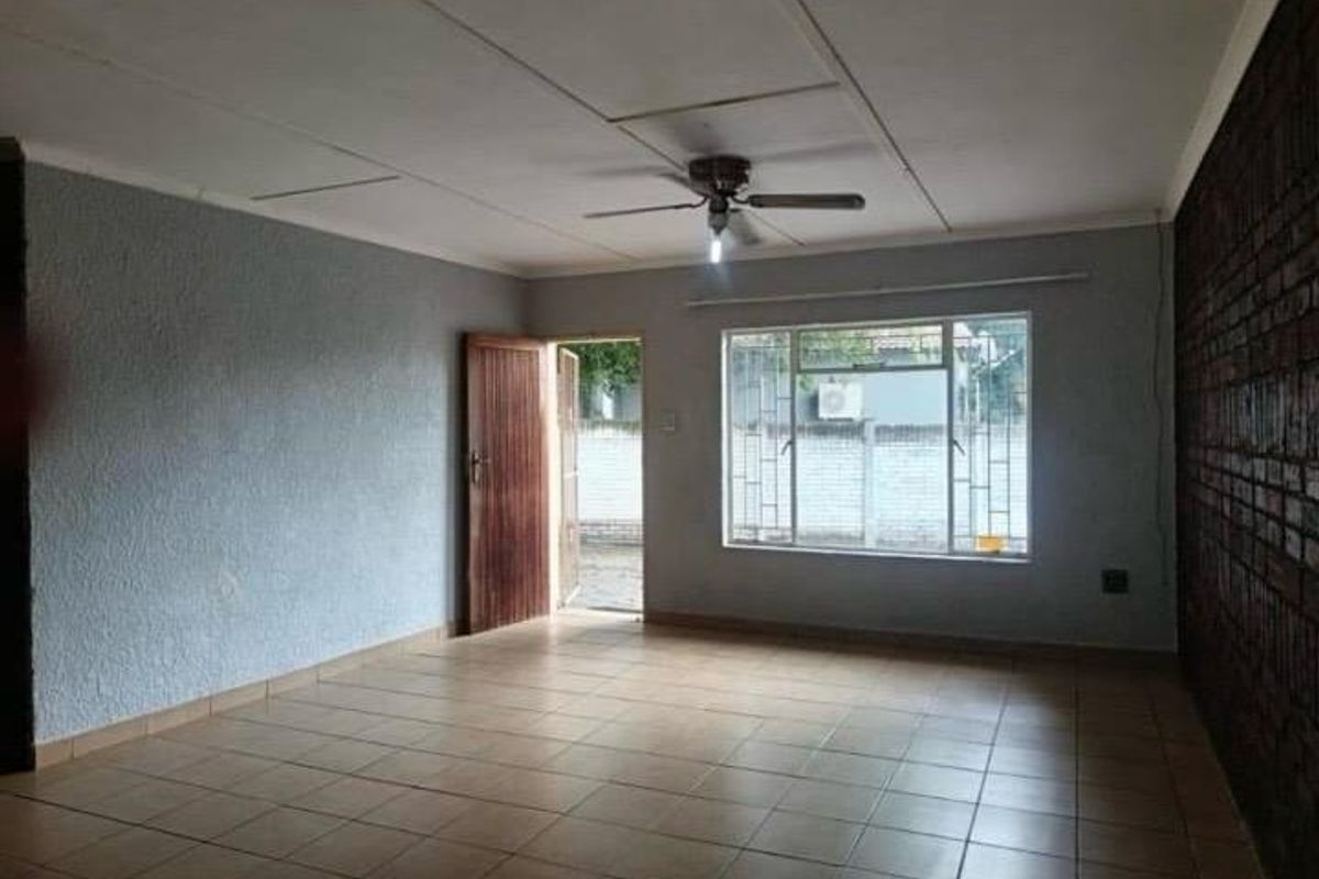 An empty living room featuring tiled flooring, an exposed brick feature wall, a ceiling fan, and security bars on the window.