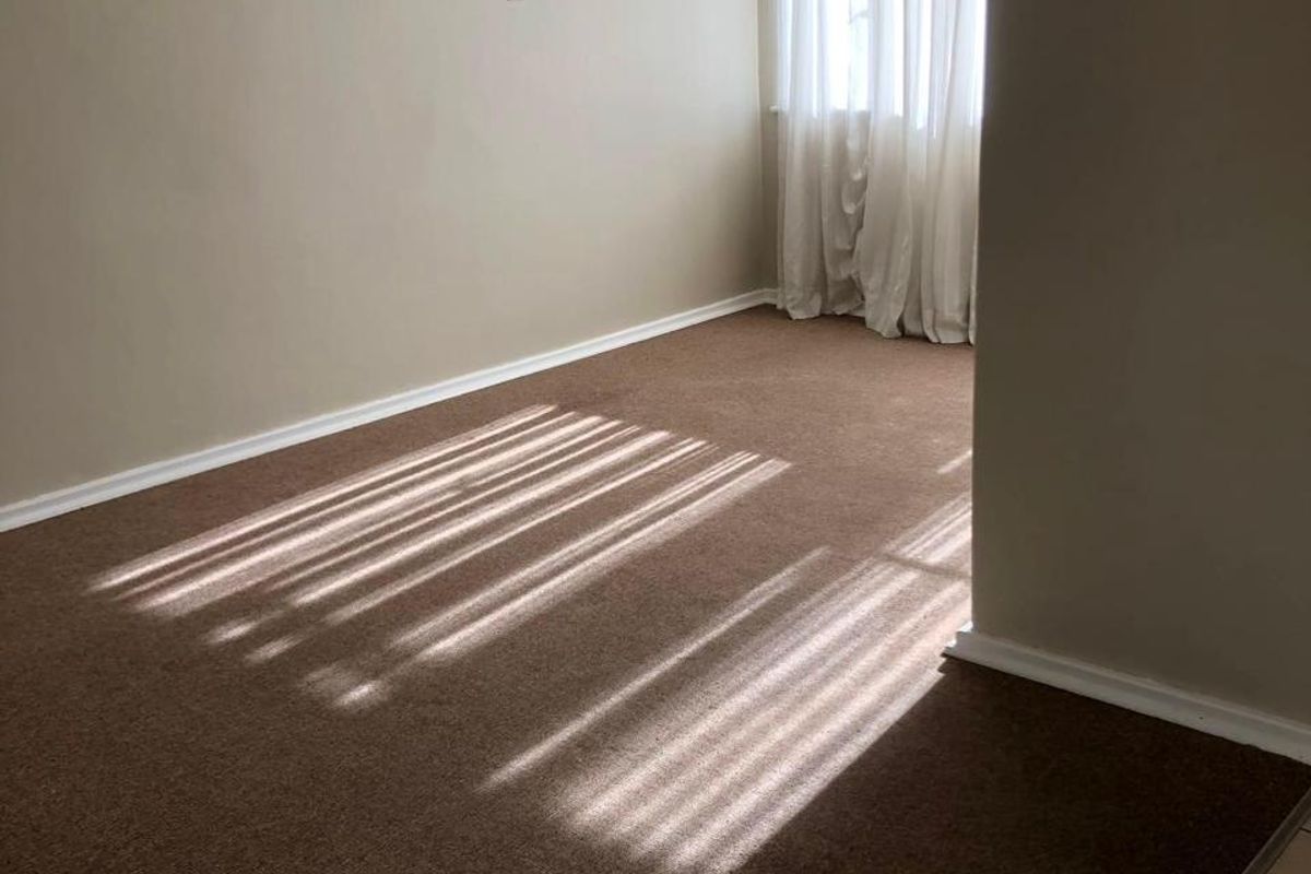 An empty bedroom featuring neutral wall paint, brown carpet flooring, and a wall-mounted light fixture with natural light filtering through window curtains.