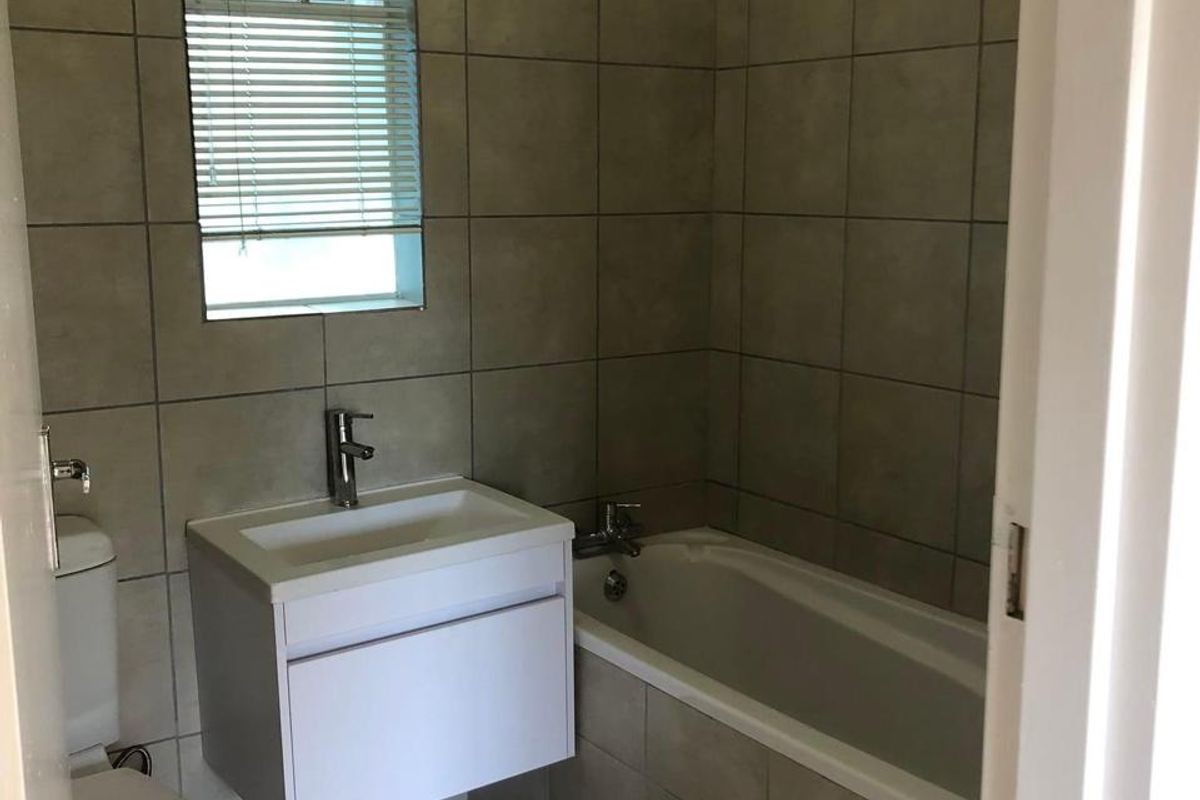 A clean, functional bathroom featuring a wall-mounted vanity, basin, toilet, and a tiled bathtub area with neutral wall and floor tiles.