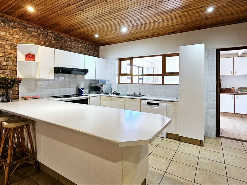 A functional kitchen featuring exposed brick walls, timber ceiling, breakfast bar, and ample built-in cabinetry, with an adjacent scullery area visible.