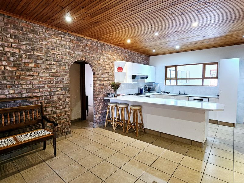 Open plan kitchen and living area featuring a rustic exposed brick wall, timber ceiling, and a functional breakfast bar with white cabinetry.