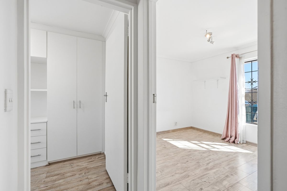 A bright, empty bedroom featuring light-toned laminate flooring, built-in storage cupboards, and a window with security bars.
