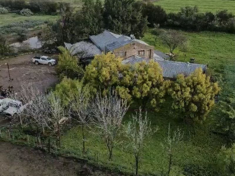 Aerial view of a rural farm property featuring a brick structure with a corrugated metal roof set within a large, partially unpaved landscape.