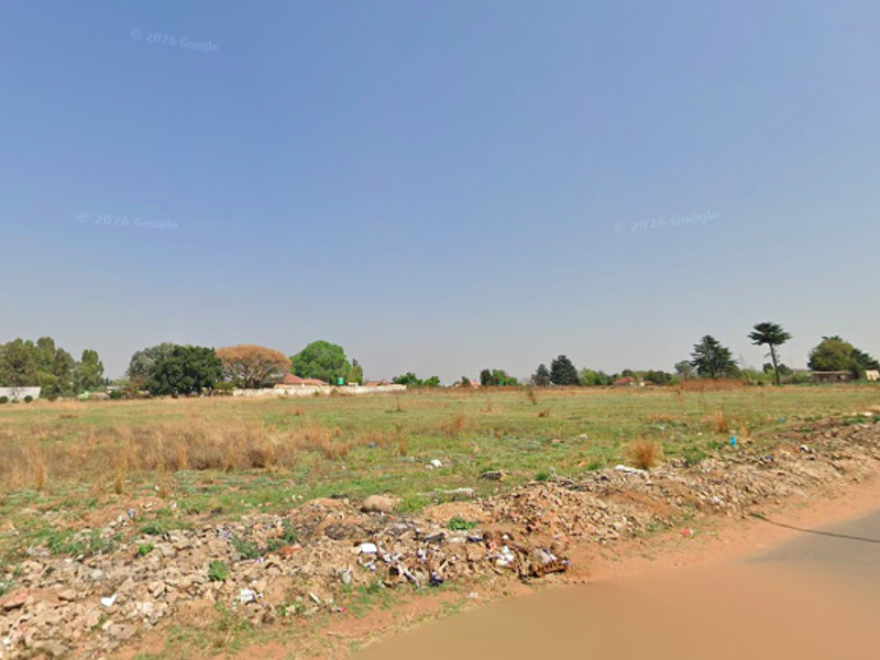 A street-level view of a vacant residential plot featuring open land with some debris along the roadside.