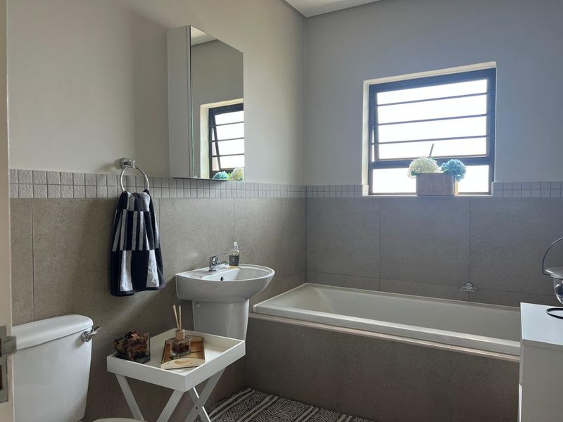 A clean, neutral-toned bathroom featuring a built-in bathtub, pedestal basin, and a mirrored medicine cabinet with natural light from a louvered window.