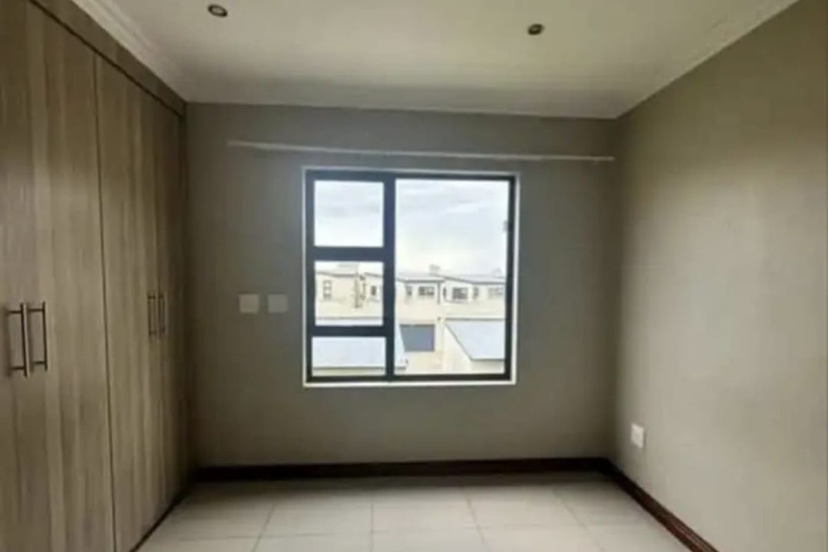 A clean, empty bedroom featuring neutral tiled flooring, built-in wooden cupboards, and a large window providing natural light.