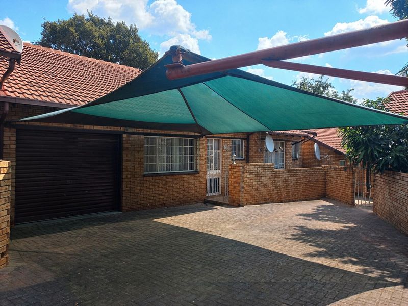 Front exterior view of a brick townhouse featuring a paved driveway, a garage, and a green shade sail structure over the parking area.