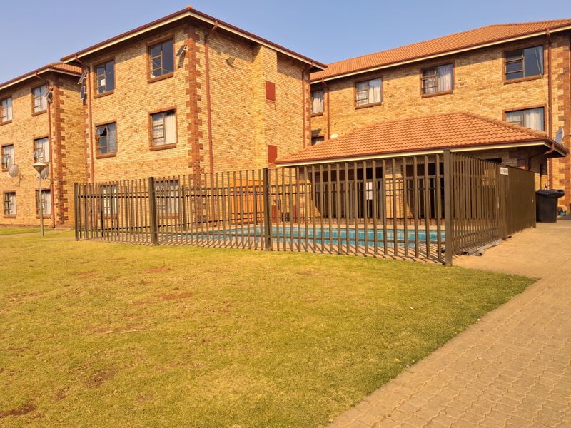 Exterior view of an apartment complex featuring a communal swimming pool enclosed by metal security fencing, set against a face brick building facade.