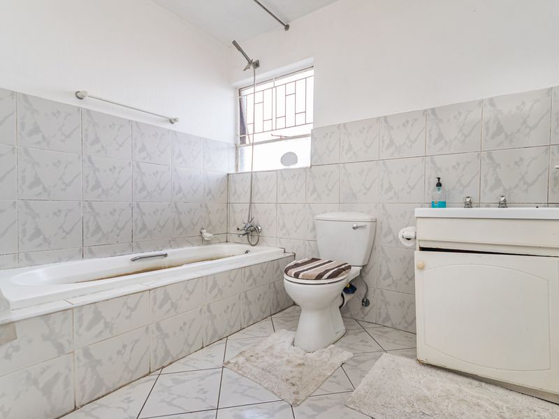 A functional bathroom featuring a bathtub, toilet, and vanity basin with tiled finishes and security bars on the window.