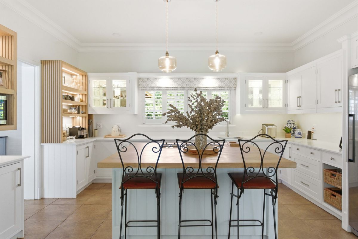 A bright, modern kitchen featuring a central island with a timber top, white cabinetry, integrated shelving, and elegant pendant lighting.