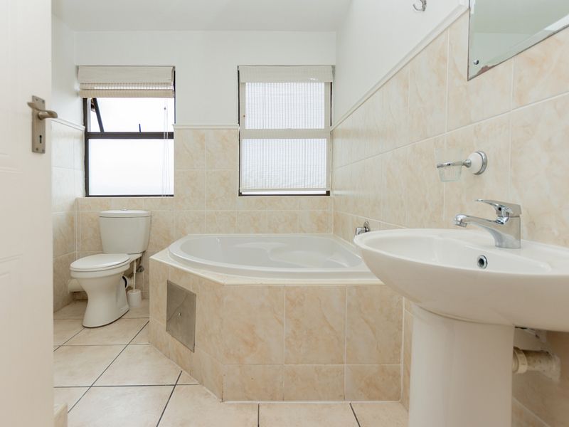 A functional bathroom featuring a corner bathtub, pedestal basin, and toilet with neutral tiled finishes and natural light from two windows.