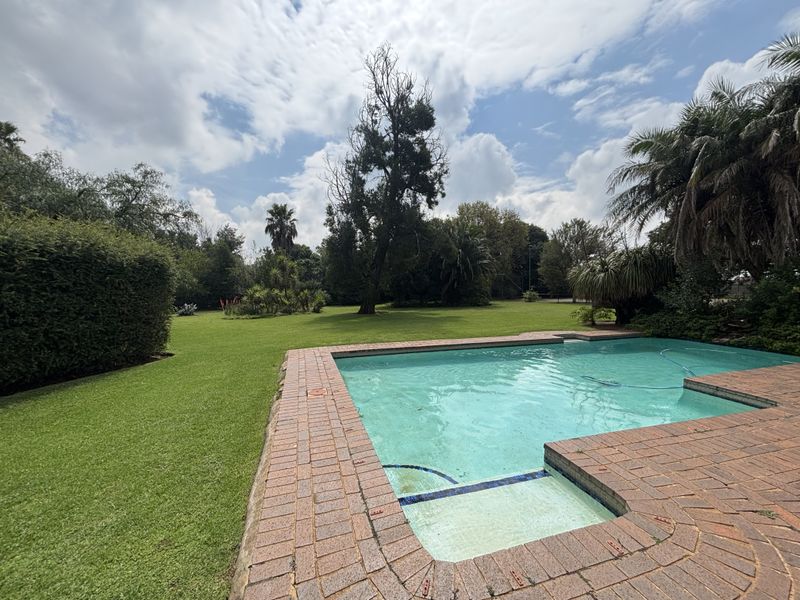 A spacious outdoor pool area featuring a swimming pool with brick coping, set within a large, well-maintained garden with mature trees.