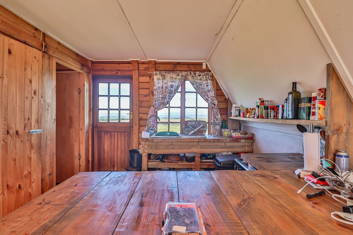 Rustic kitchen area featuring timber wall cladding, wooden countertops, open shelving, and a sink, with a view through the timber-framed door and window.