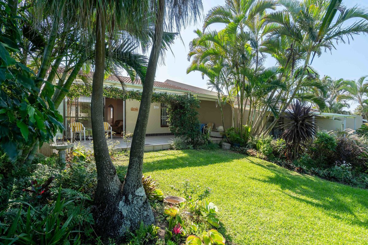 A well-maintained rear garden featuring a paved patio area, lush tropical landscaping, and direct access to the home through sliding glass doors.