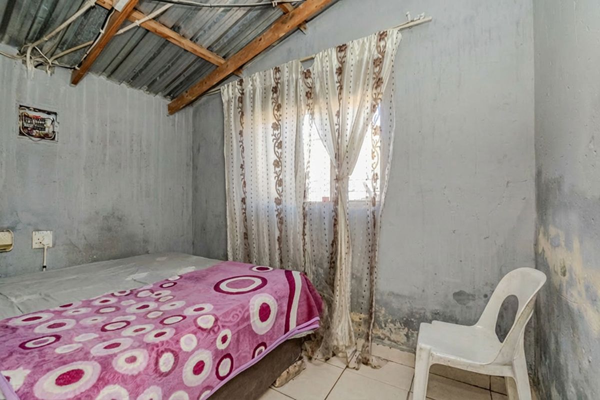 A bedroom featuring exposed timber rafters, corrugated metal roofing, and tiled flooring, currently showing significant signs of wear and wall surface deterioration.
