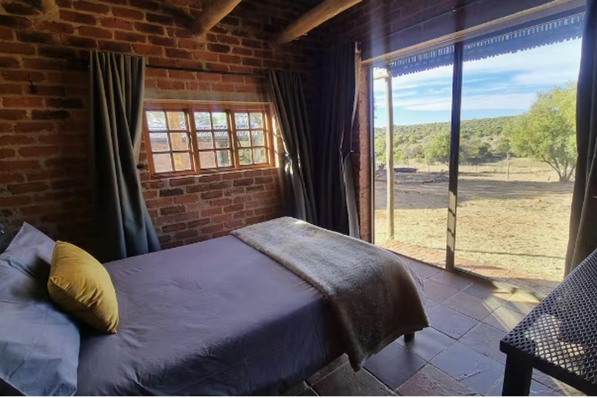 Rustic bedroom featuring exposed brick walls, timber ceiling beams, tiled flooring, and a large sliding glass door offering scenic farm views.