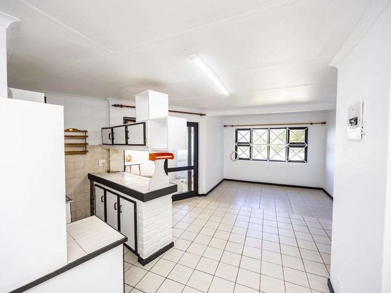An open-plan kitchen and living area featuring tiled flooring, built-in cabinetry, and large windows with security bars, offering a blank canvas for renovation.