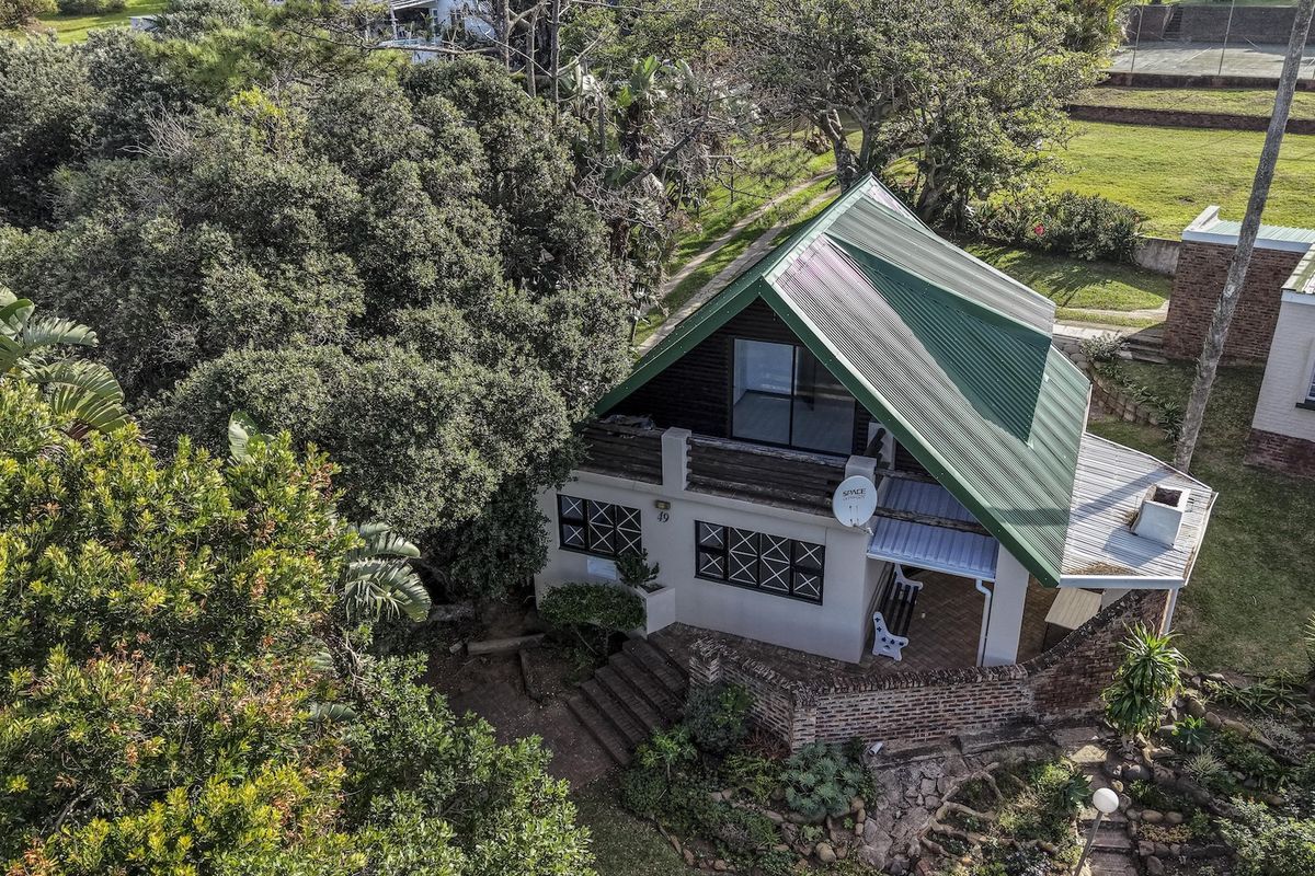 Aerial view of a detached house featuring a pitched metal roof, brick exterior walls, and a balcony, set within a lush, tree-lined garden.