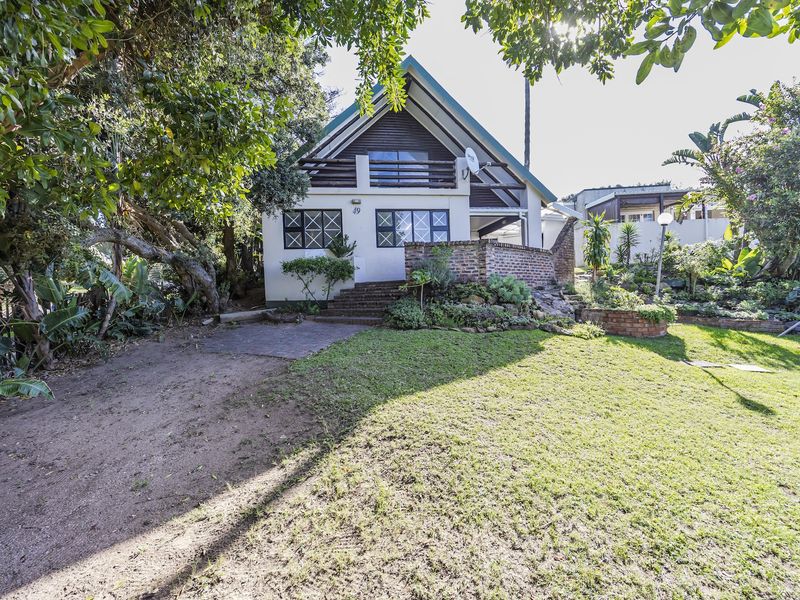 A charming A-frame house featuring a front balcony, brick retaining wall, and established garden setting with mature trees.